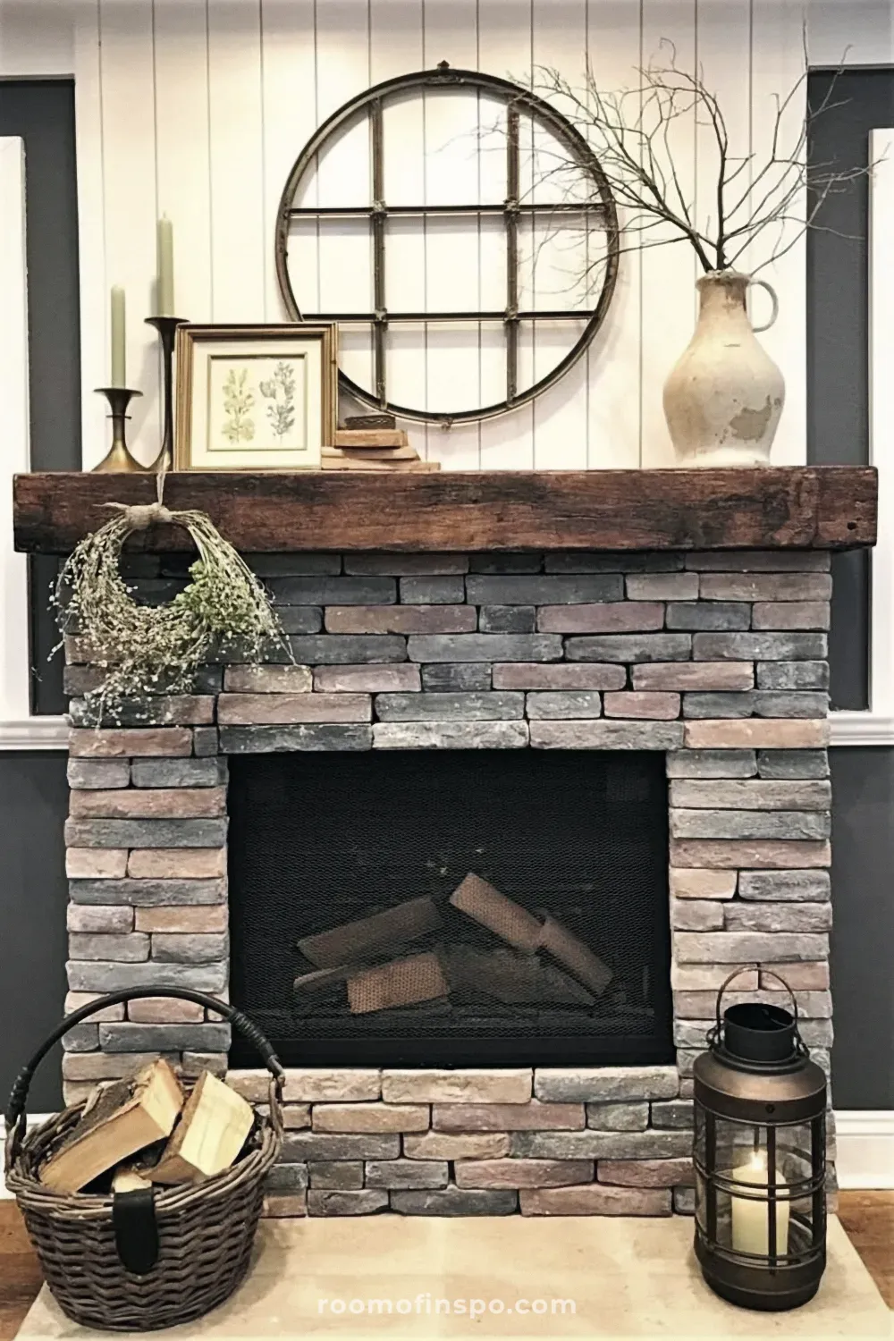 Farmhouse stacked stone fireplace with a dark wood mantel decorated with a round window frame, a vase of branches, and candles.