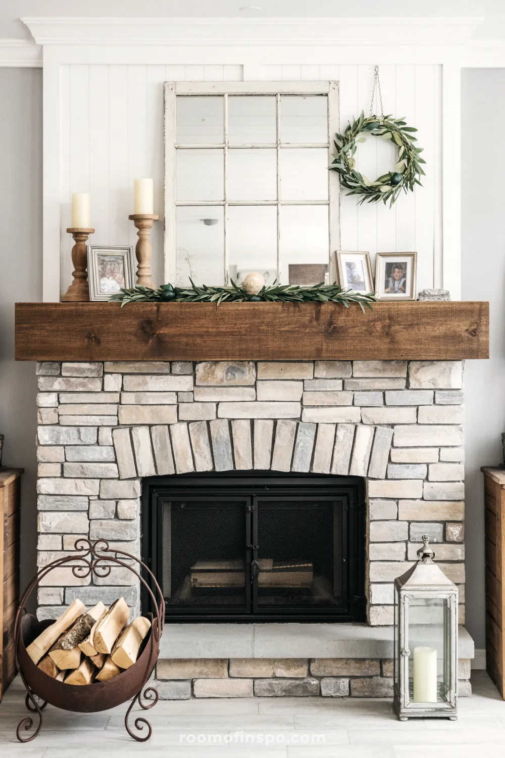 Farmhouse fireplace with a light stacked stone surround and a thick wood mantel decorated with a salvaged window mirror and a simple green wreath.