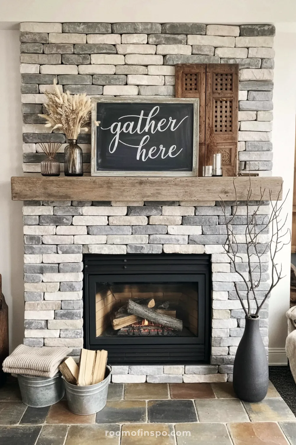 Rustic stone fireplace with a thick wood mantel displaying a "gather here" sign, dried grasses, and a carved wood accent.