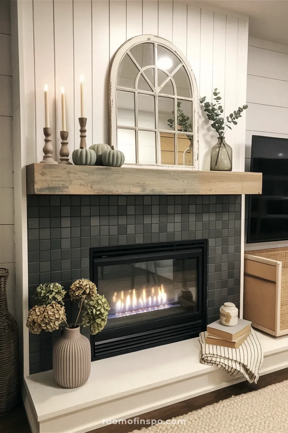 Farmhouse fireplace with dark tile and a distressed wood mantel displaying an arched window mirror, taper candles, and small blue pumpkins.