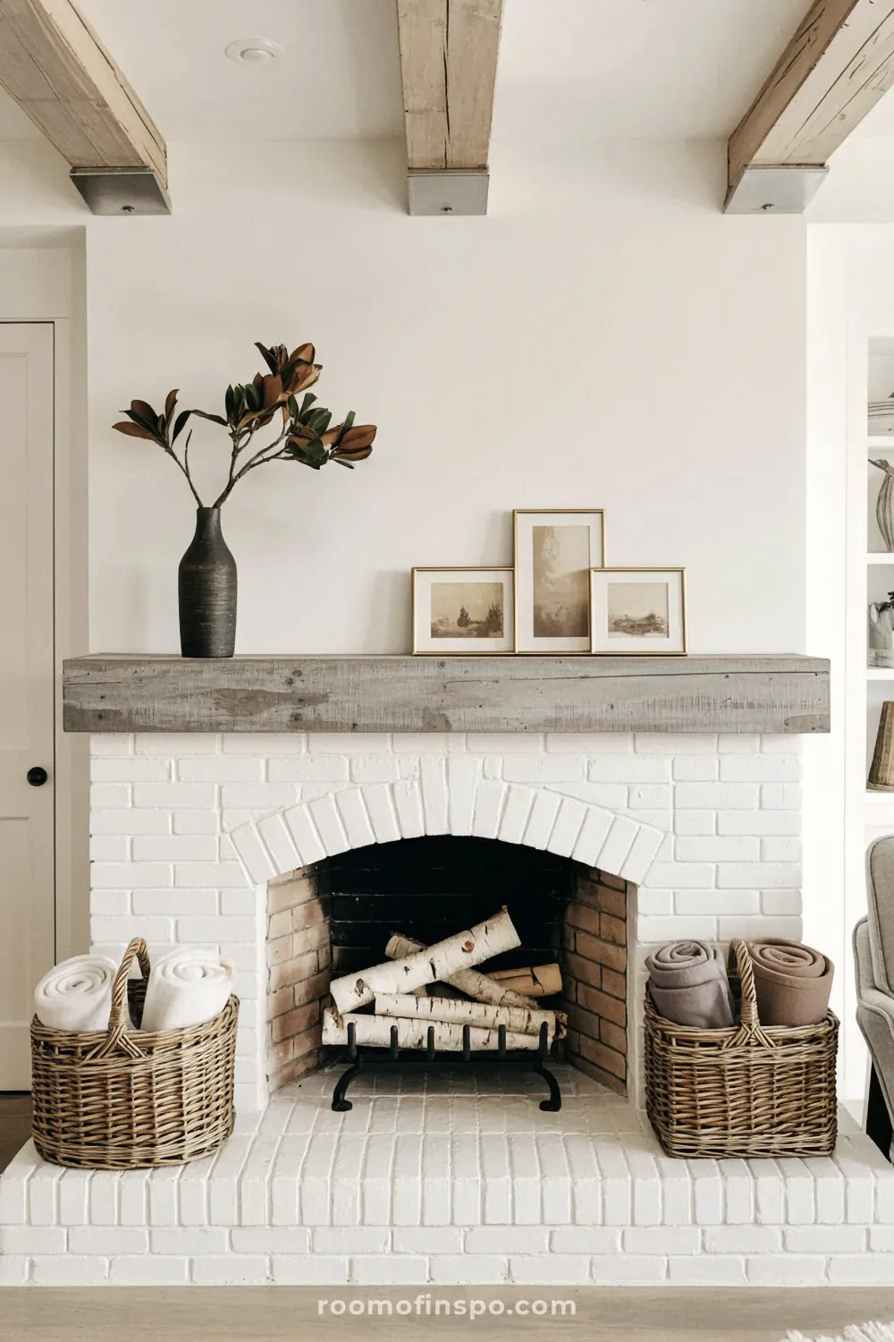 Farmhouse fireplace with white painted brick and a rustic gray wood mantel displaying a vase of leaves and three small framed landscape pictures.