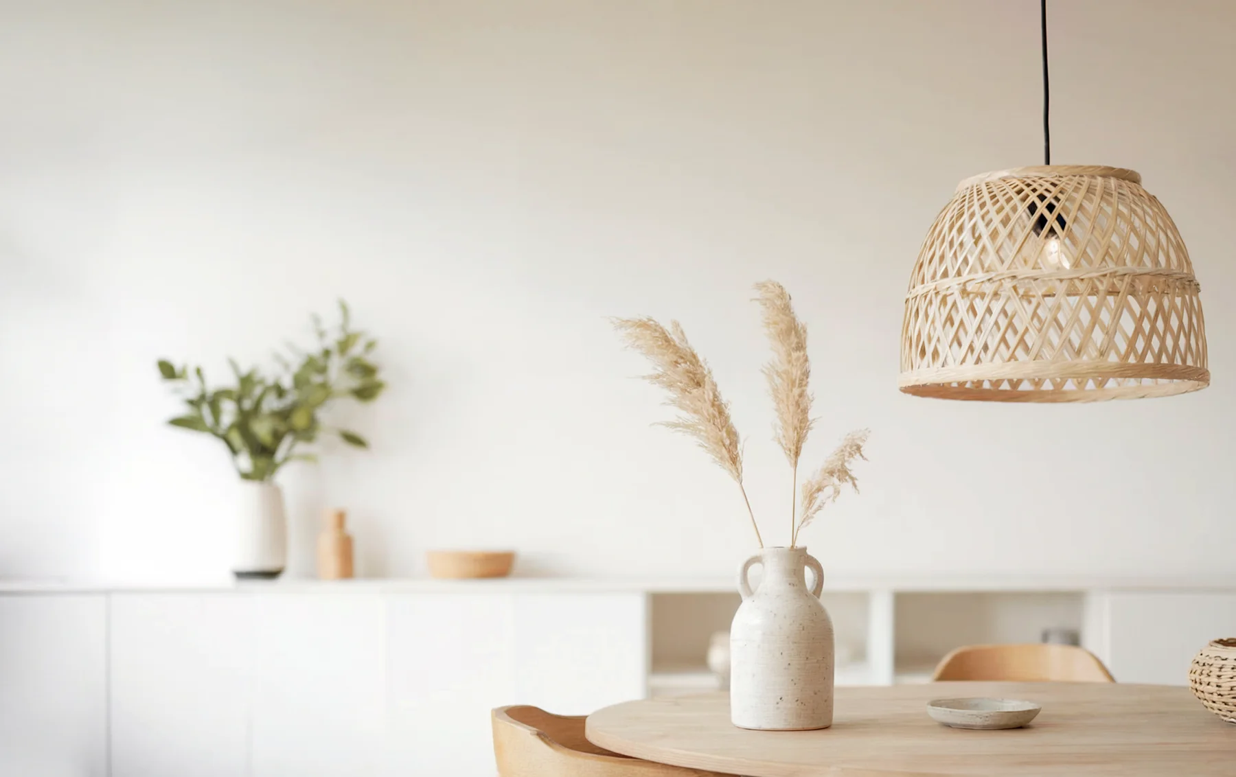 A bright, minimalist dining room features a round wooden table with a white vase holding dried pampas grass, a woven pendant light, and a glimpse of greenery in a vase on a white sideboard.