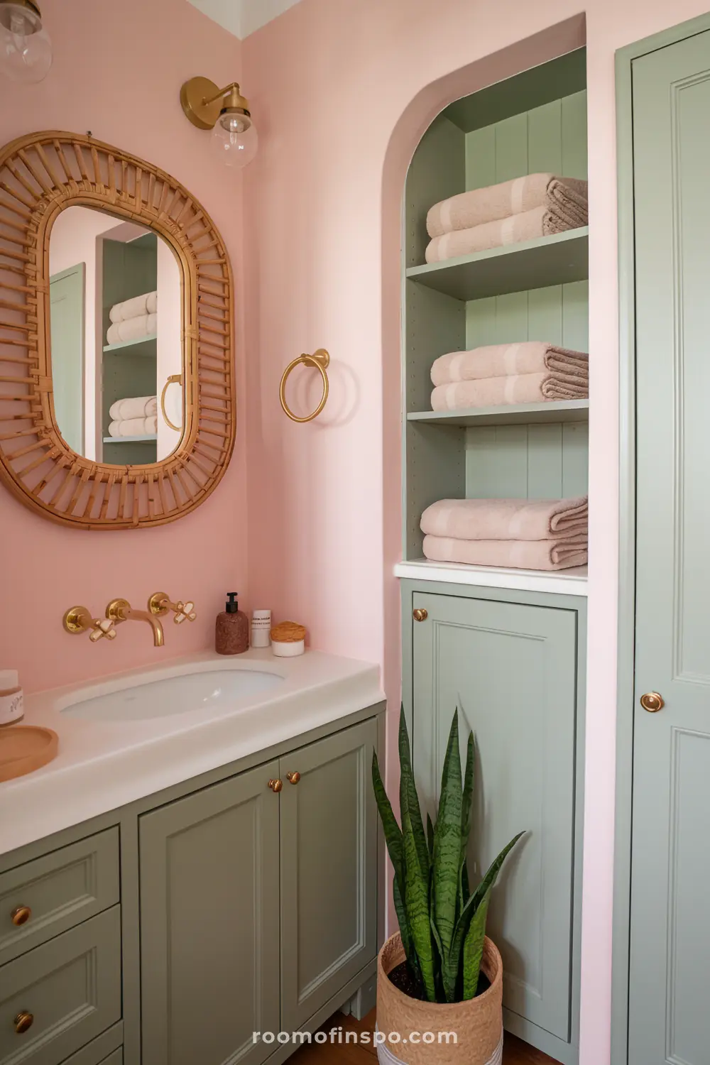 A bohemian bathroom with blush pink walls, sage green cabinets, and a rattan-framed mirror.