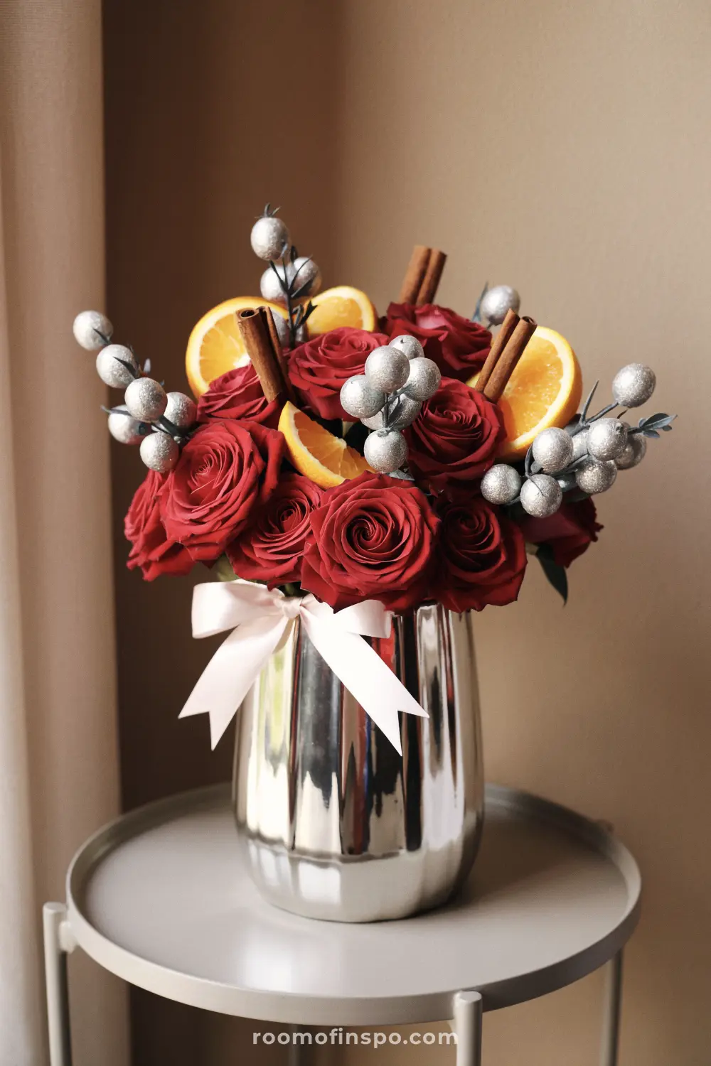 A striking and classy Christmas centerpiece of red roses, silver berries, cinnamon sticks, and orange slices in a shiny silver vase.