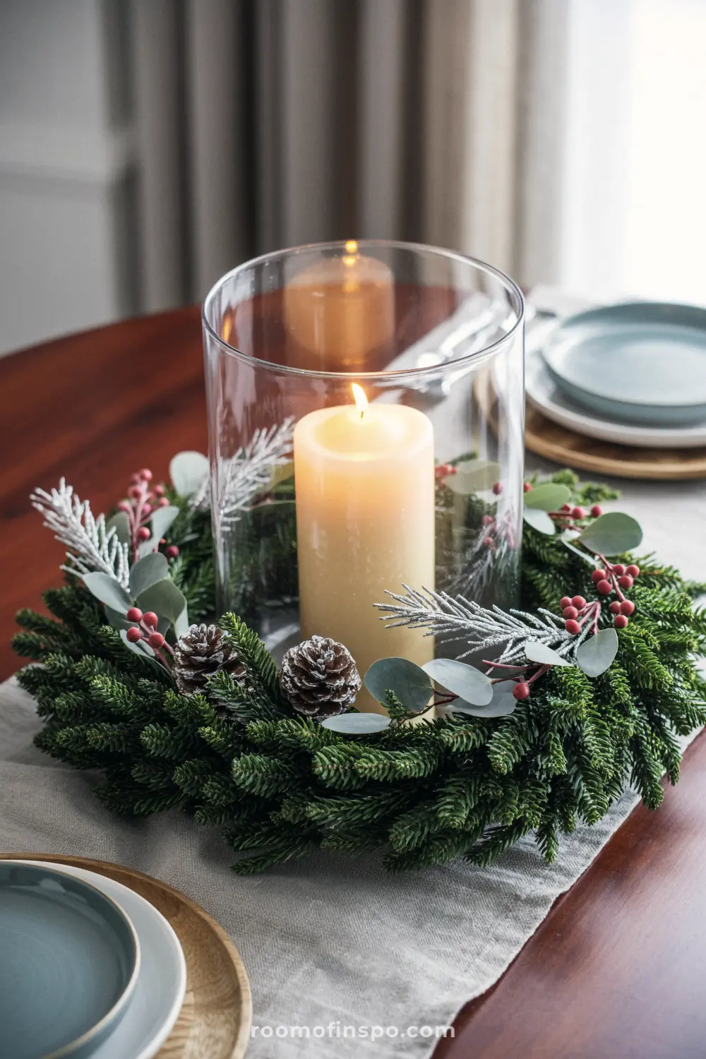 A thick pine wreath with berries and eucalyptus encircles a tall candle in a glass, creating a minimalist Christmas centerpiece.