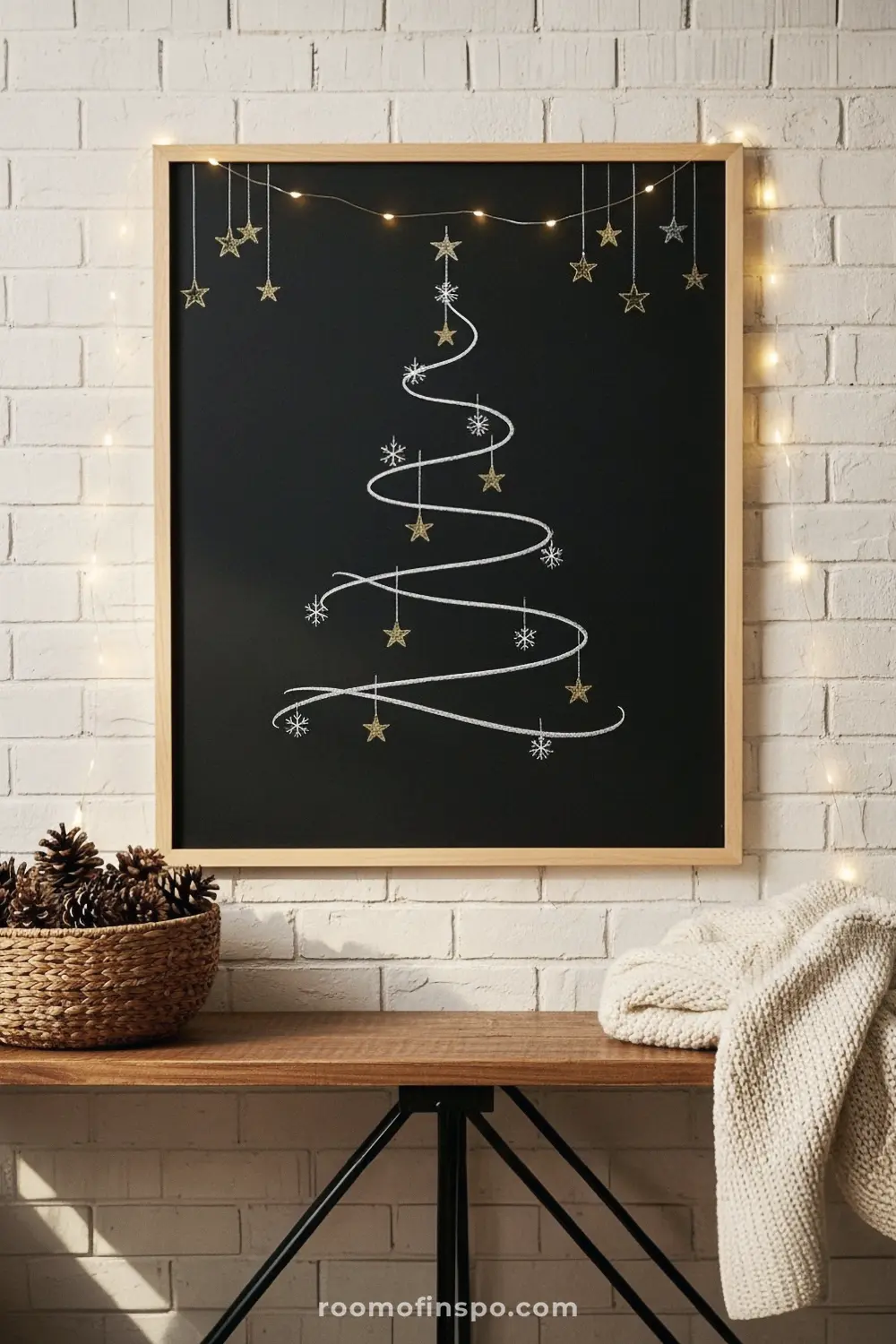 A minimalist Christmas tree is drawn on a framed chalkboard with a swirling line, star ornaments, and snowflakes, illuminated by string lights.