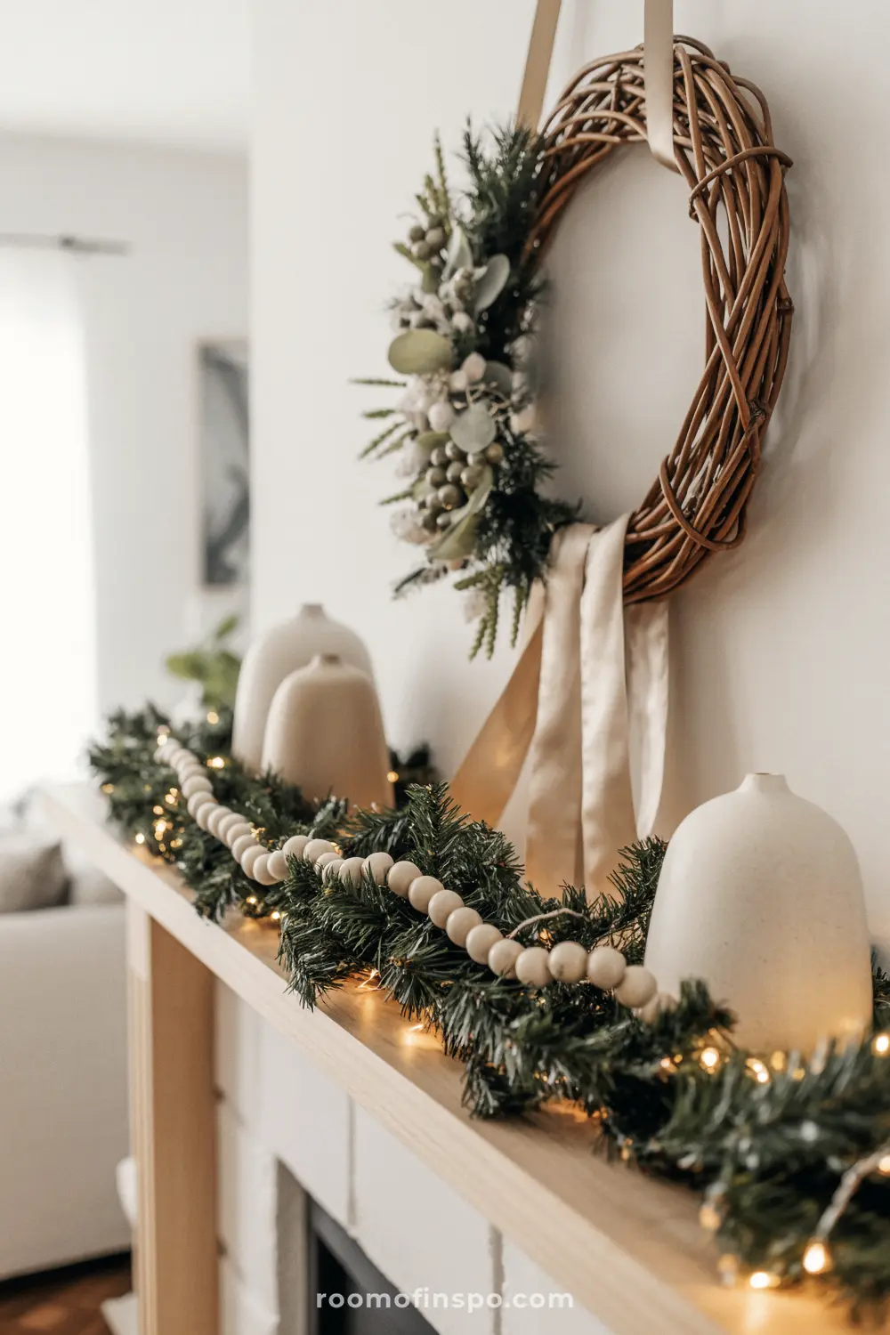 A rustic, neutral Christmas mantel featuring a simple rattan wreath, a lush garland with string lights, and modern white vases.