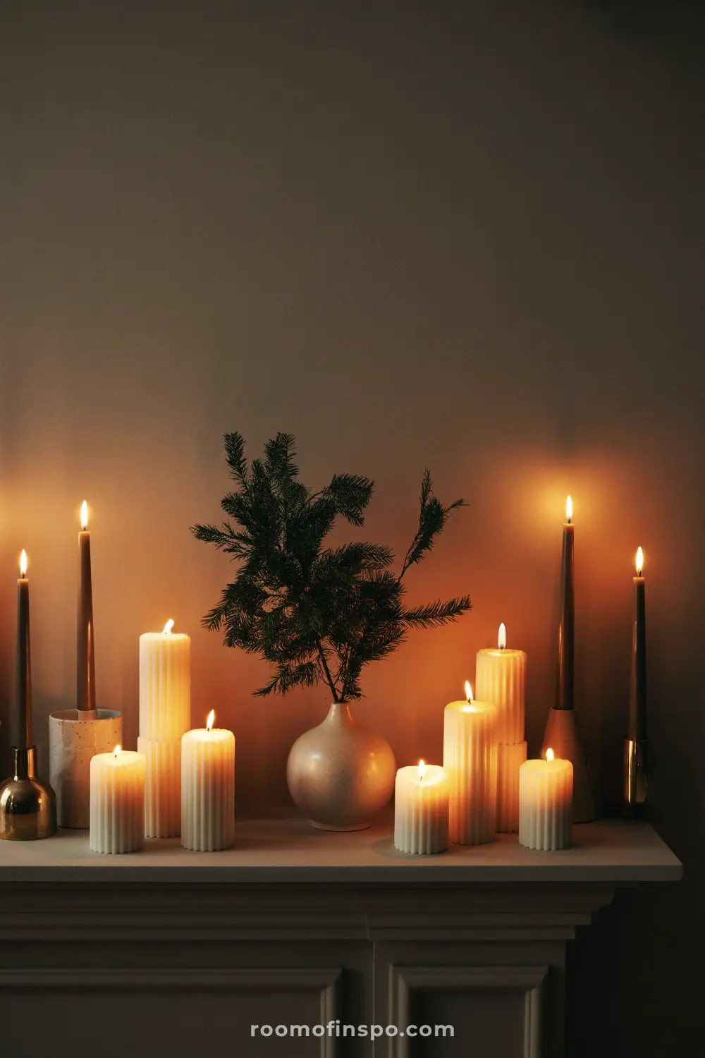 A cozy Christmas mantel glowing with multiple pillar and taper candles surrounding a small evergreen branch in a vase.