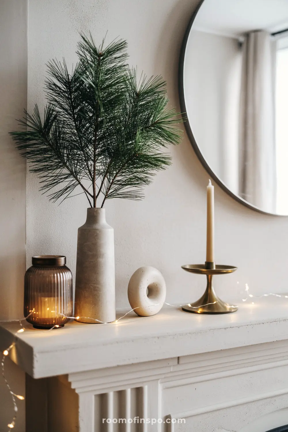 A light mantel is adorned with a concrete vase of pine, a brass candle holder, and lights for minimalist Christmas cheer.