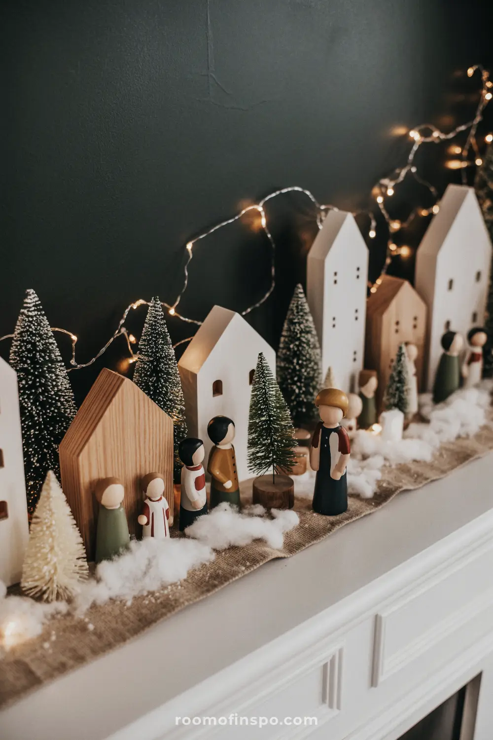 A delightful Christmas mantel village with wooden houses, bottle brush trees, and simple figures on a burlap runner.