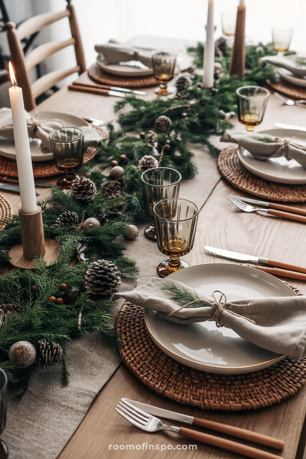 Rustic Christmas table setting on natural wood, featuring woven chargers, linen napkins, amber glassware, and a centerpiece of pinecones and evergreen garland.