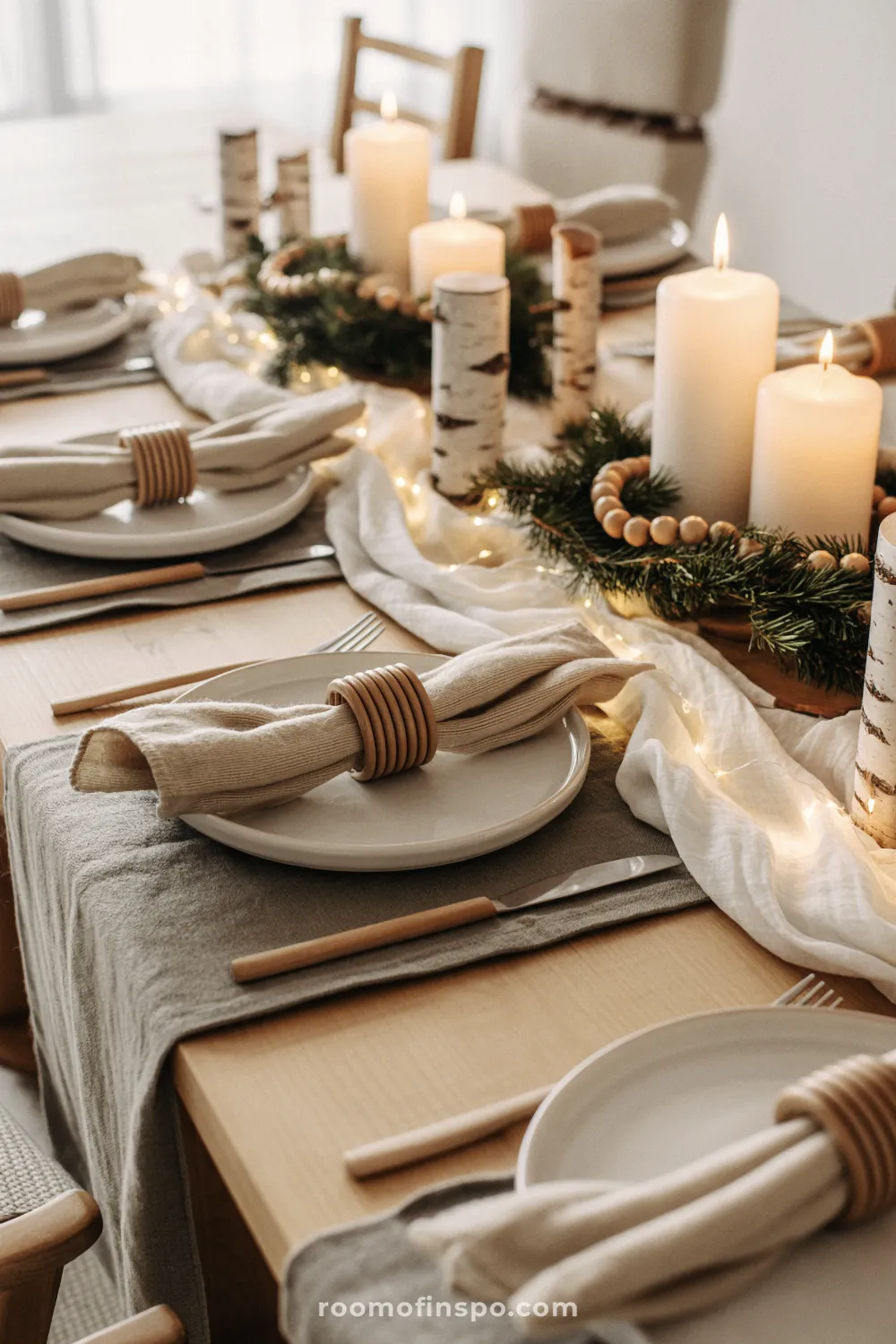 Cozy Nordic Christmas table setting featuring white plates, wooden napkin rings, birch-wrapped candles, and warm fairy lights on a sheer white runner.