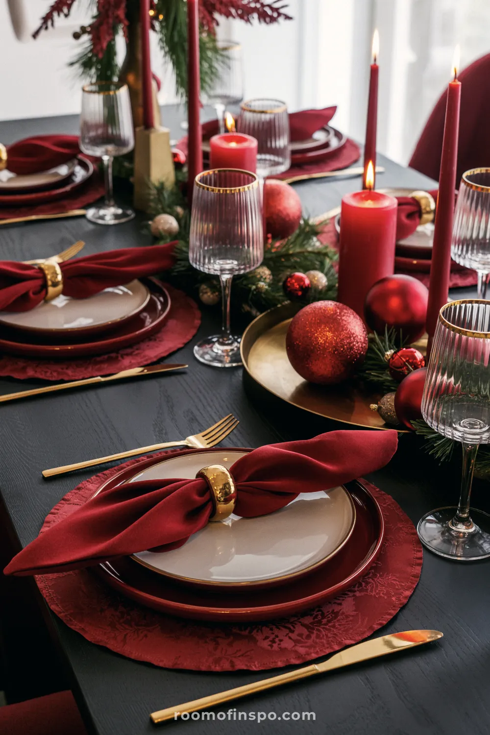 Dramatic Christmas table setting in deep red and gold, featuring patterned placemats, tall candles, ribbed glassware, and glittering festive ornaments.