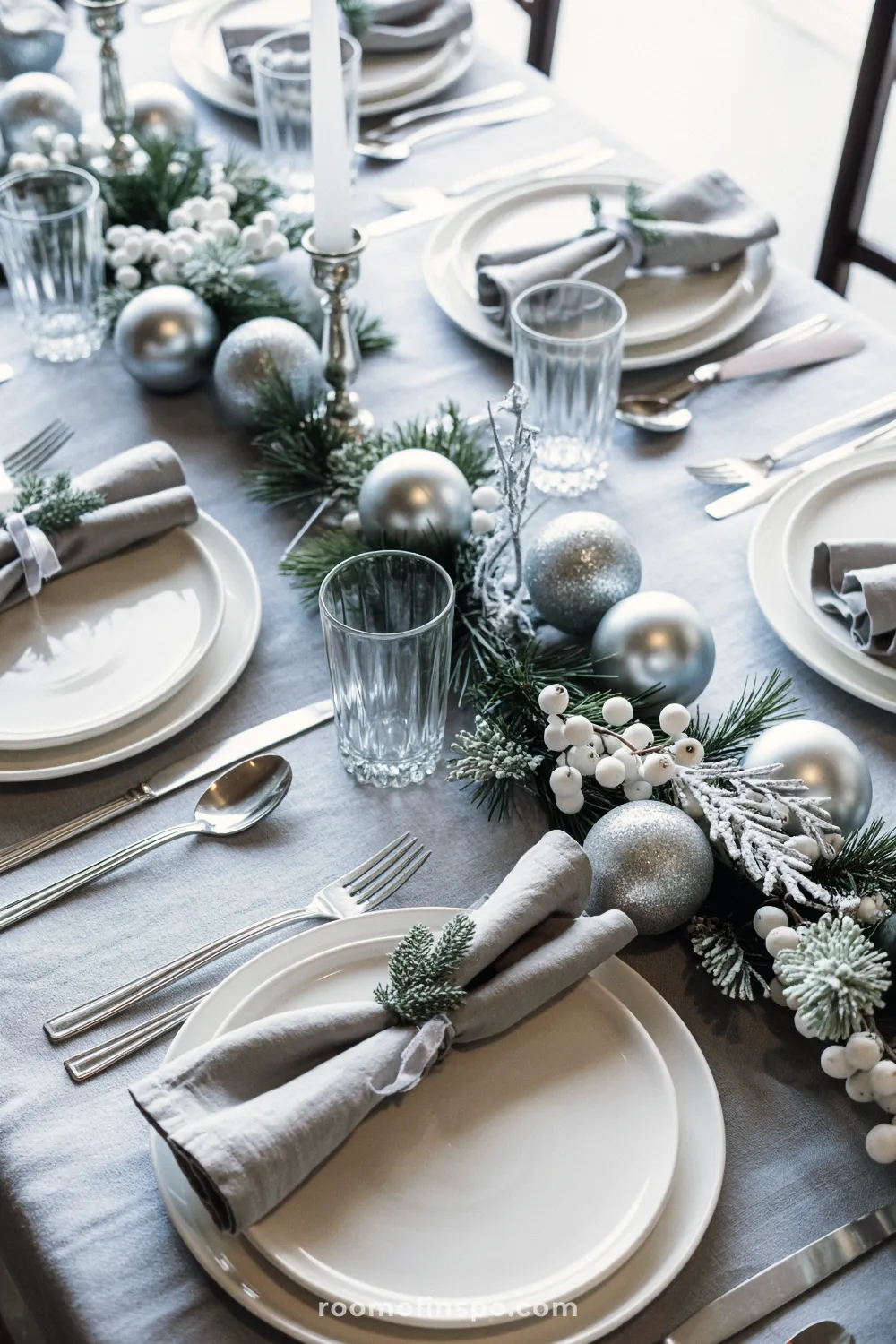 Snowy white and silver Christmas table setting on a light gray tablecloth with linen napkins, simple white plates, and frosted winter greenery garland.