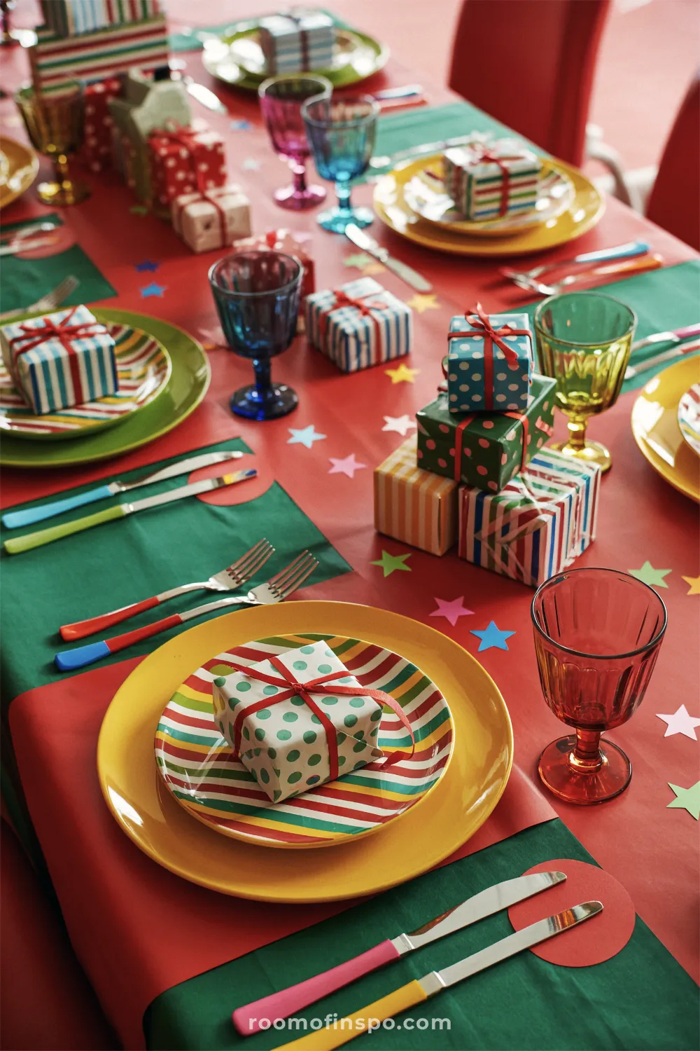 Playful and bright Christmas table setting with solid red and green placemats, striped plates, colorful goblet glassware, and small wrapped gifts.