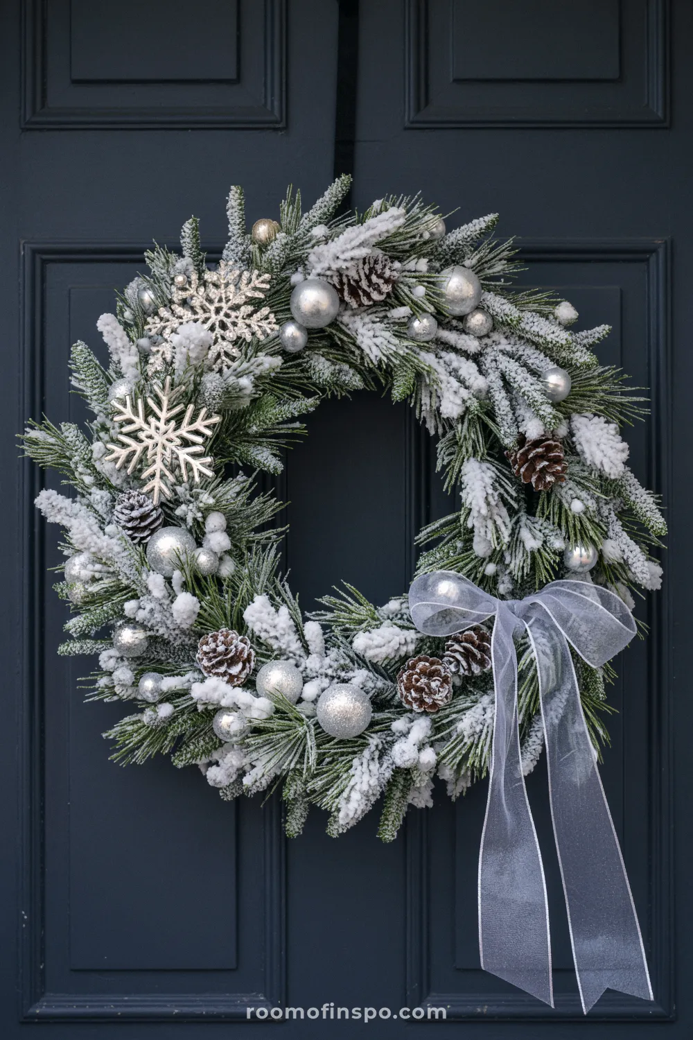 A snowy and classy Christmas wreath with silver ornaments, pinecones, snowflakes, and a sheer white ribbon on a dark door.