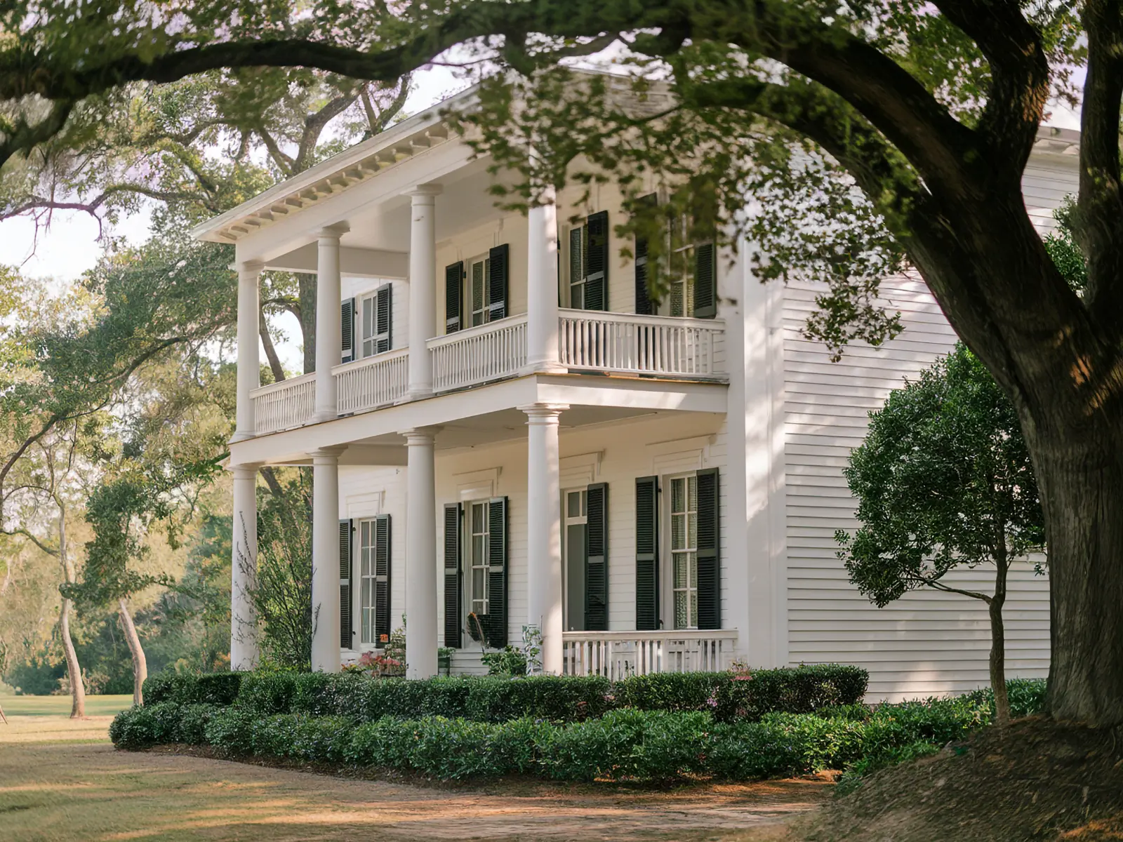 A large classic southern white house with a wraparound porch and black shutters, surrounded by lush trees and greenery.