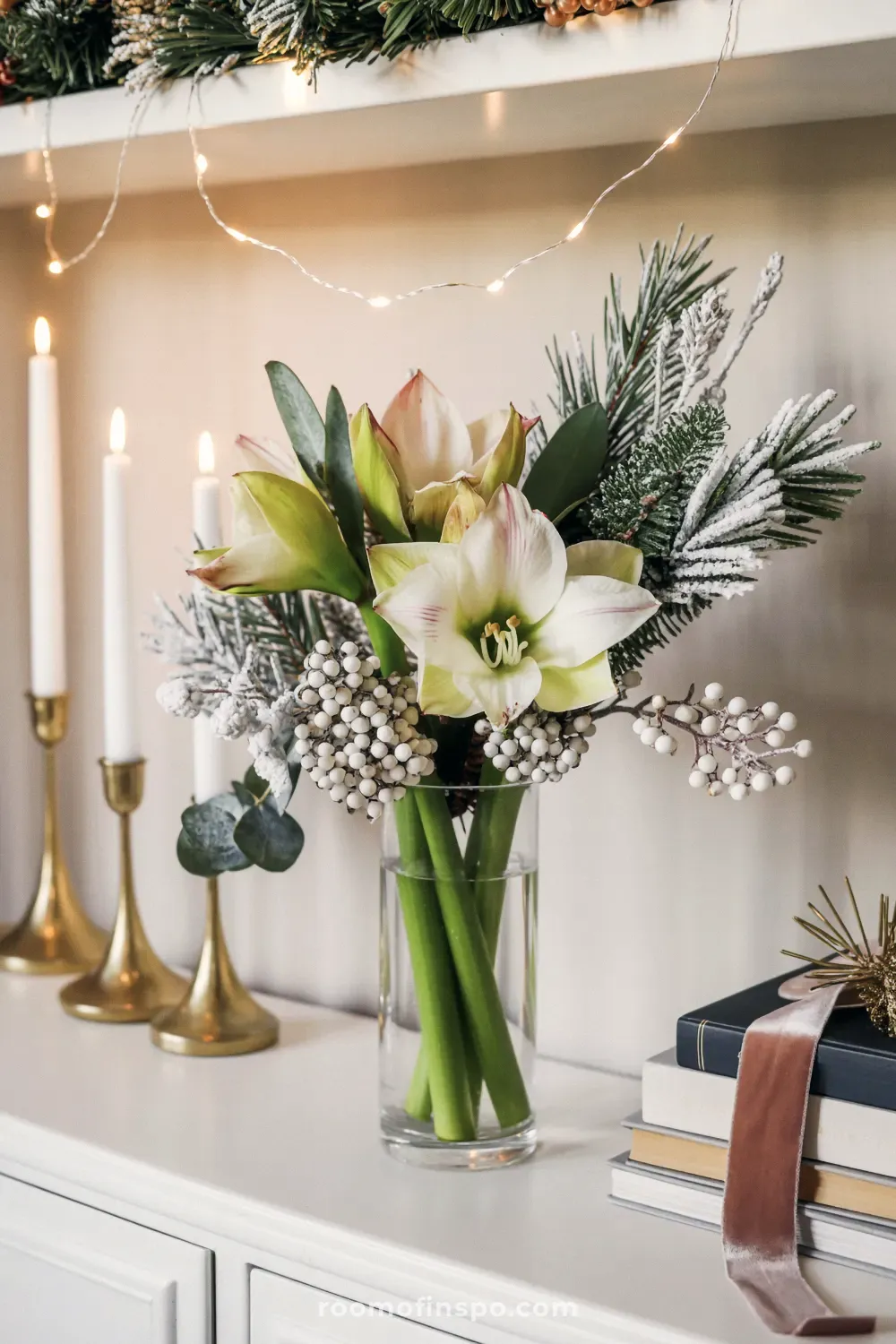 A delicate and classy Christmas floral arrangement featuring white amaryllis, flocked pine, and winter berries next to brass candlesticks.
