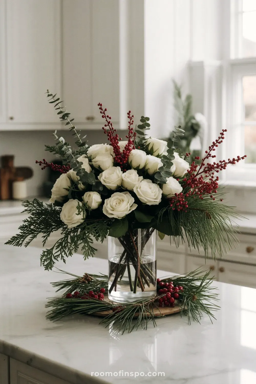 A gorgeous and classy Christmas arrangement of white roses, red hypericum berries, and fresh evergreen in a clear vase on a counter.