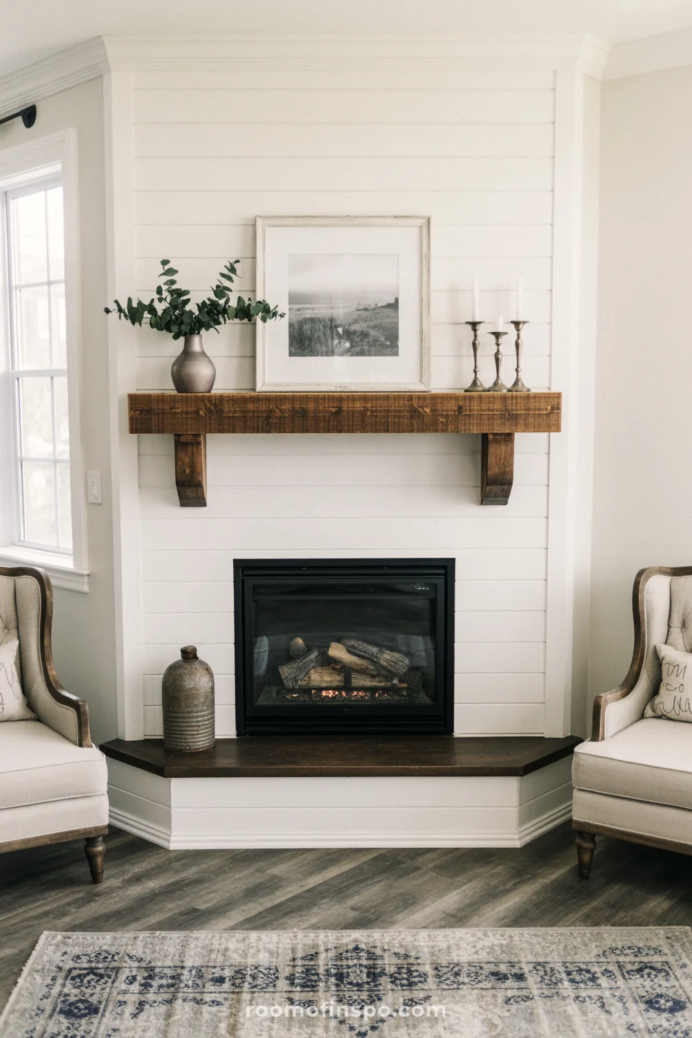 White shiplap corner fireplace with wood mantel, beige armchair, light wood floors, and muted patterned rug.