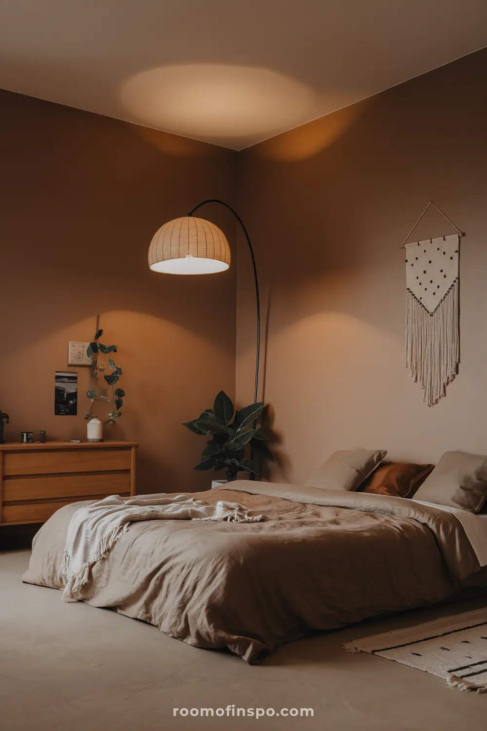 A cozy bedroom with warm lighting, featuring a bed with beige linens, a wooden nightstand with decor items, a large potted plant, a hanging lamp, and a macrame wall hanging.