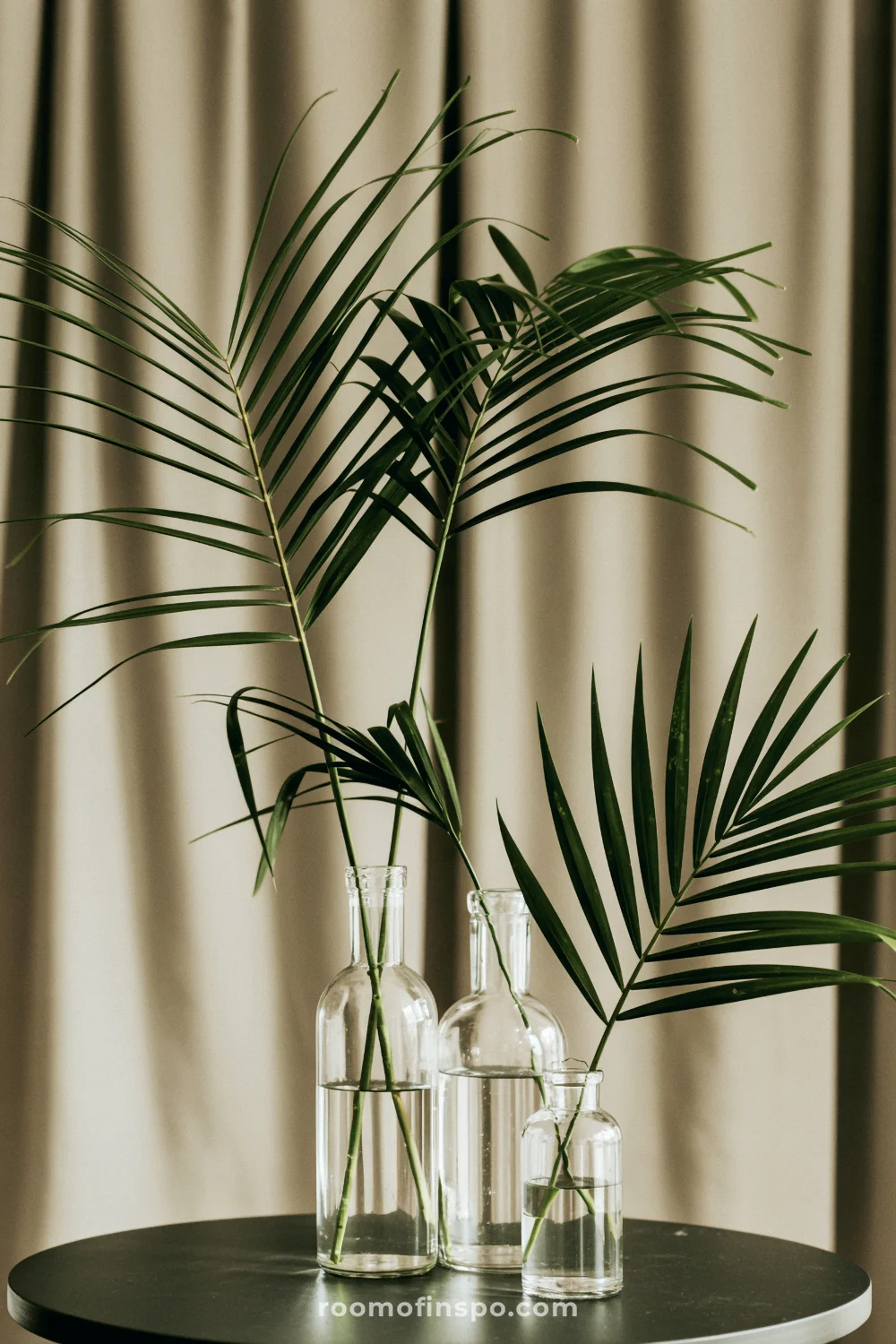 Palm fronds in clear glass bottles on a dark tabletop with a neutral background.