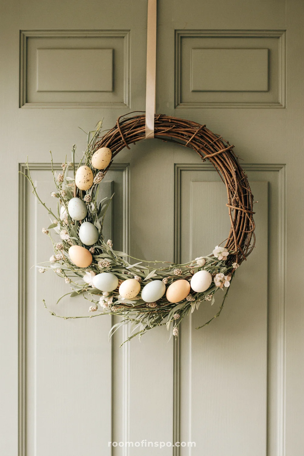 A minimalist Easter wreath with pastel eggs and dried florals on a sage-green door.