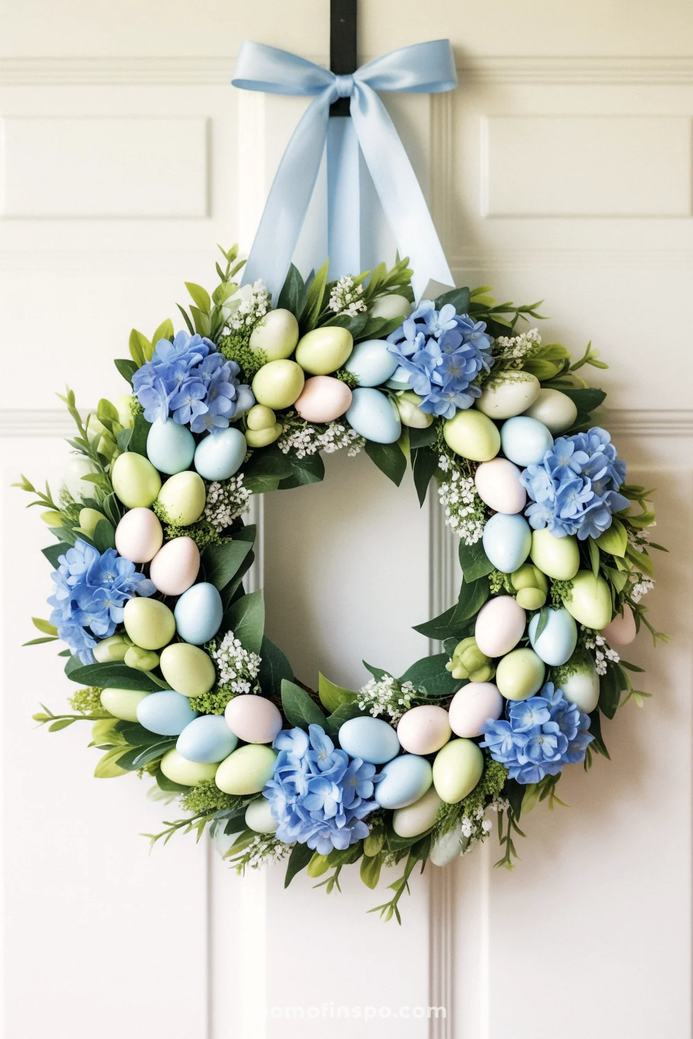 A pastel Easter wreath with eggs and hydrangea-style blooms on a white door.