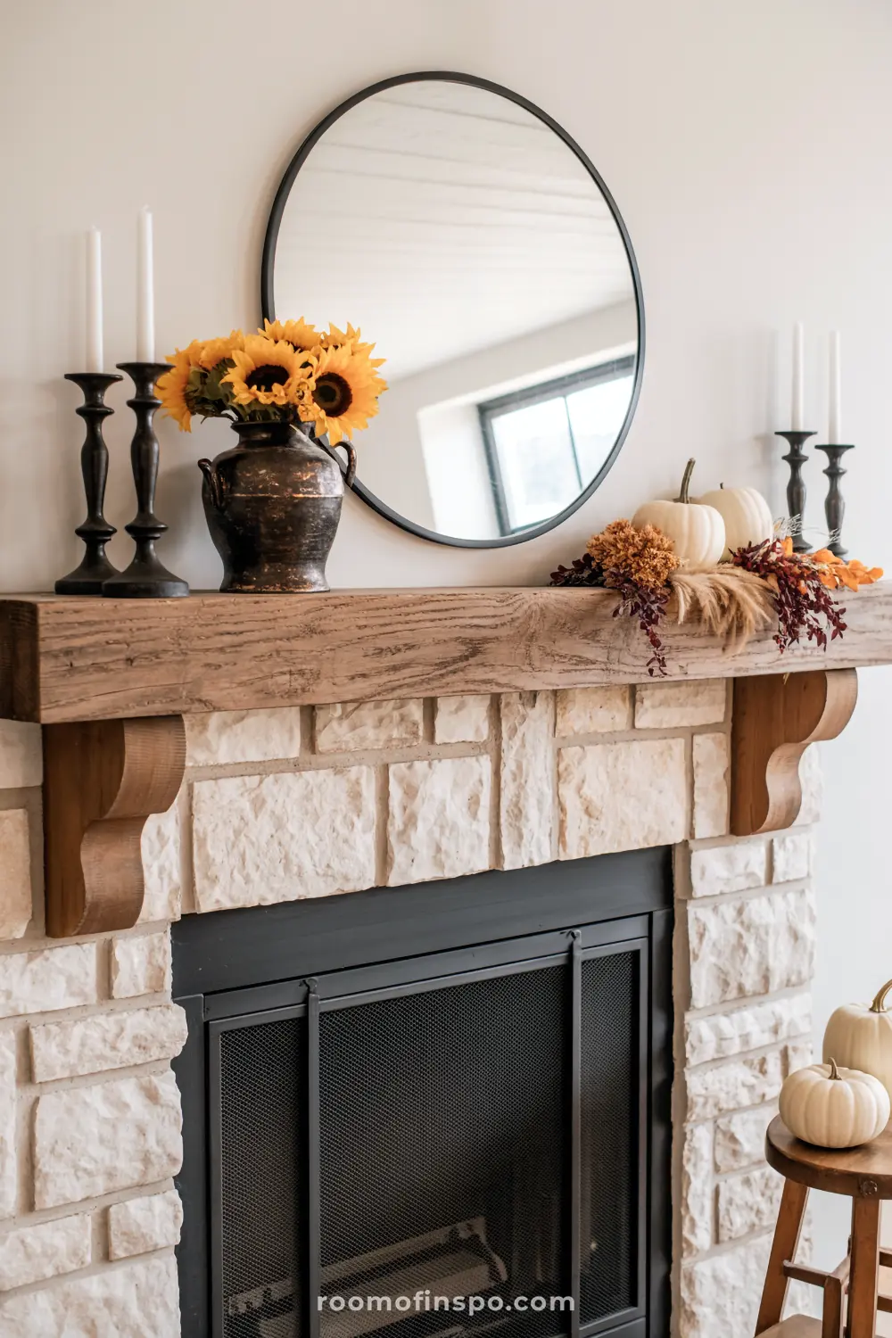 A cozy farmhouse mantel decorated for fall with sunflowers, white pumpkins, candles, and a round mirror over a stone fireplace.
