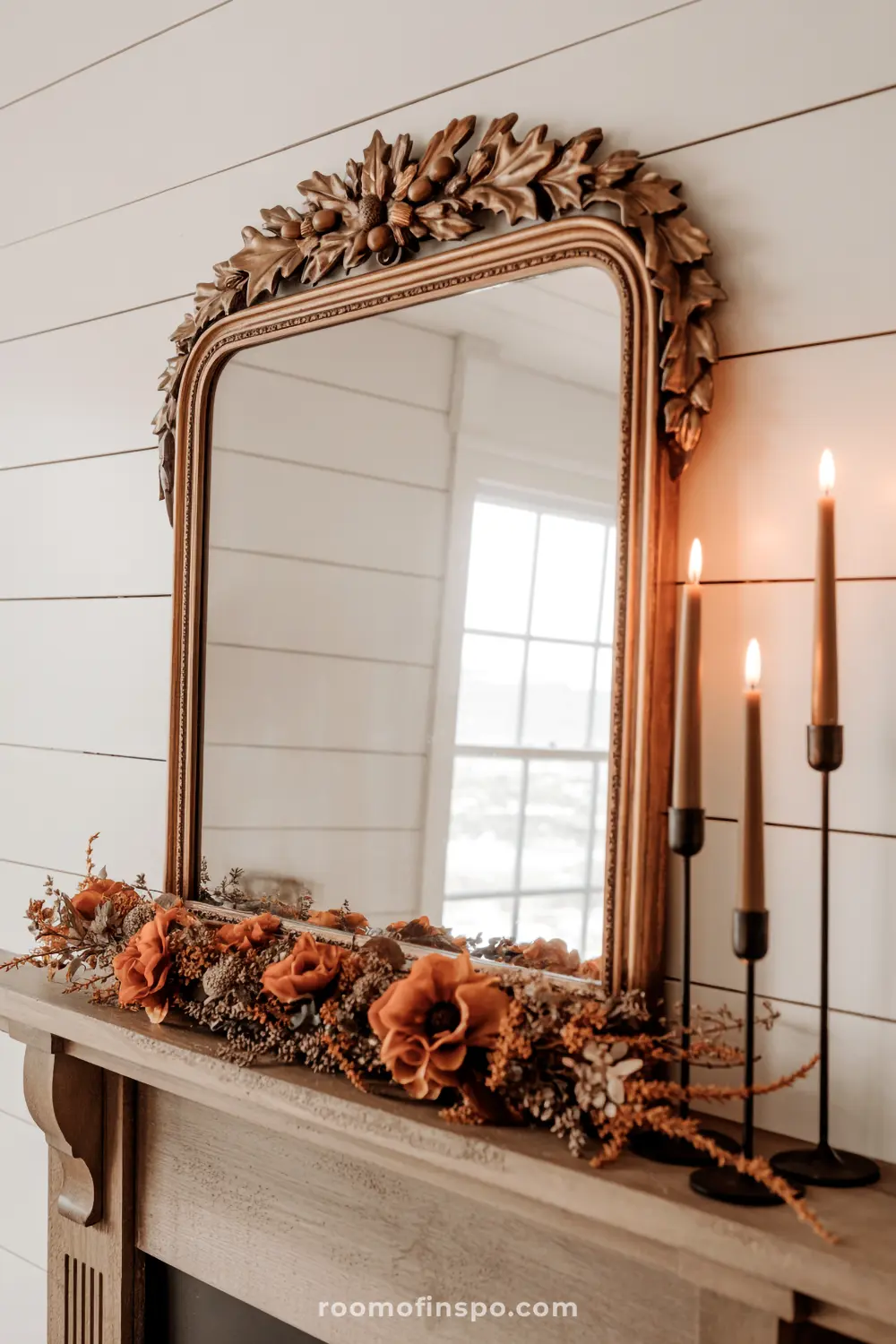 An elegant fall mantel features a decorative mirror with an acorn motif, accented by a garland of orange blooms and flickering candles.