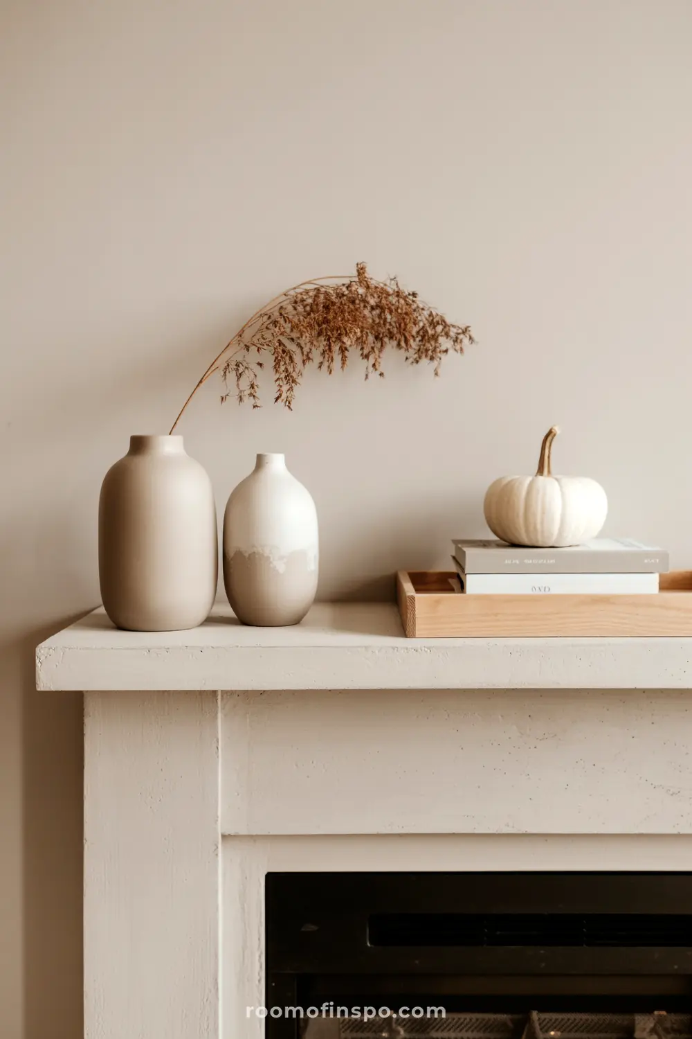 A neutral-toned fall mantel displays a white pumpkin on books, two vases with dried foliage, and a wooden tray.
