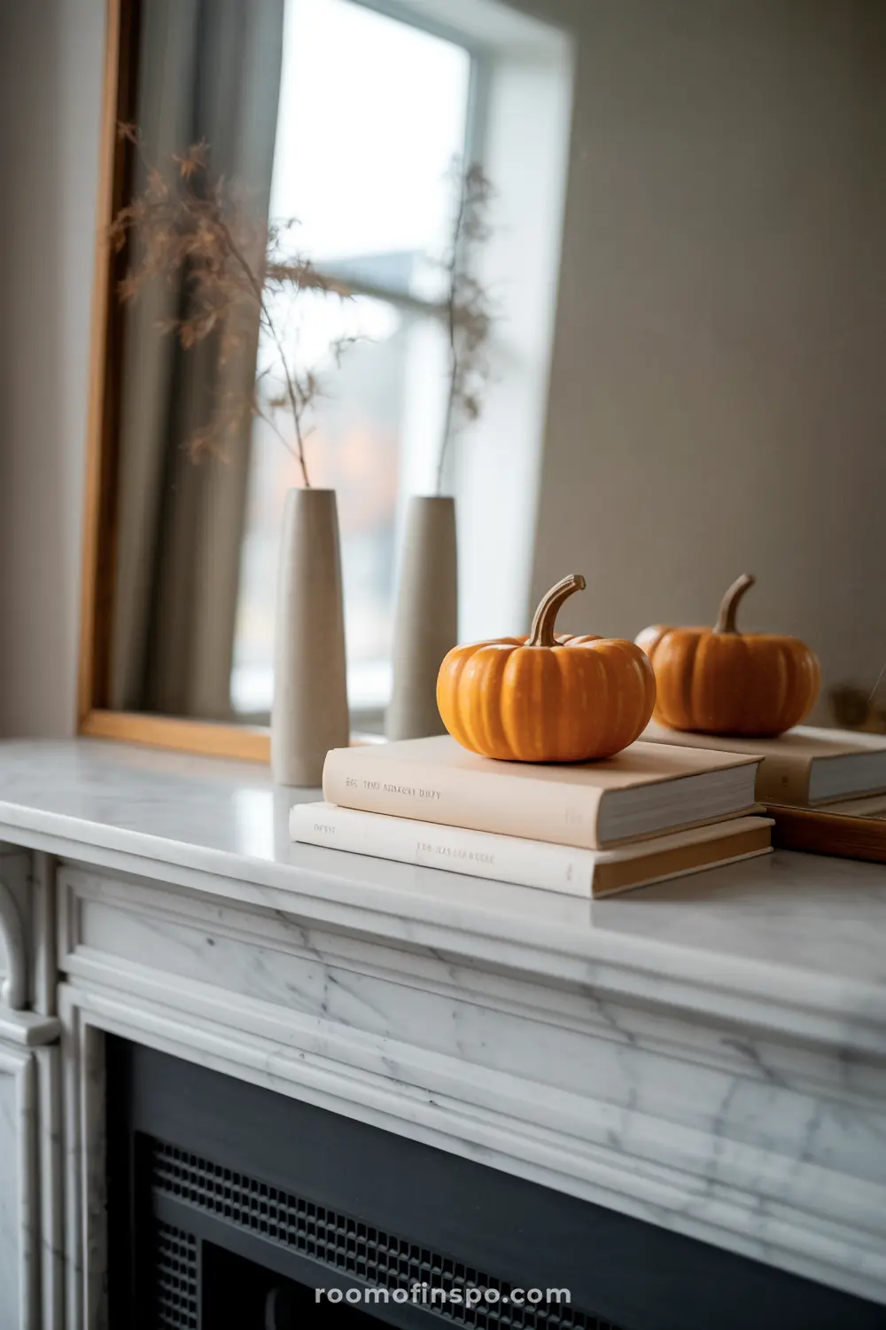 A minimalist fall mantel features two small orange pumpkins atop books, flanked by slender vases with dried branches, reflecting in a mirror.