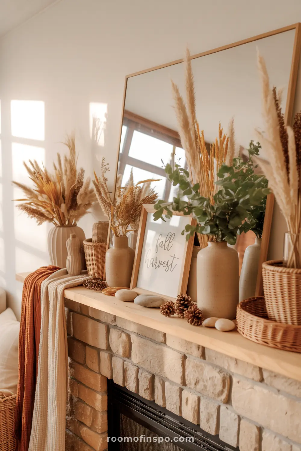 A beautifully styled fall mantel features various dried grasses, eucalyptus, pinecones, and a "fall harvest" sign, with a mirror reflecting the room.