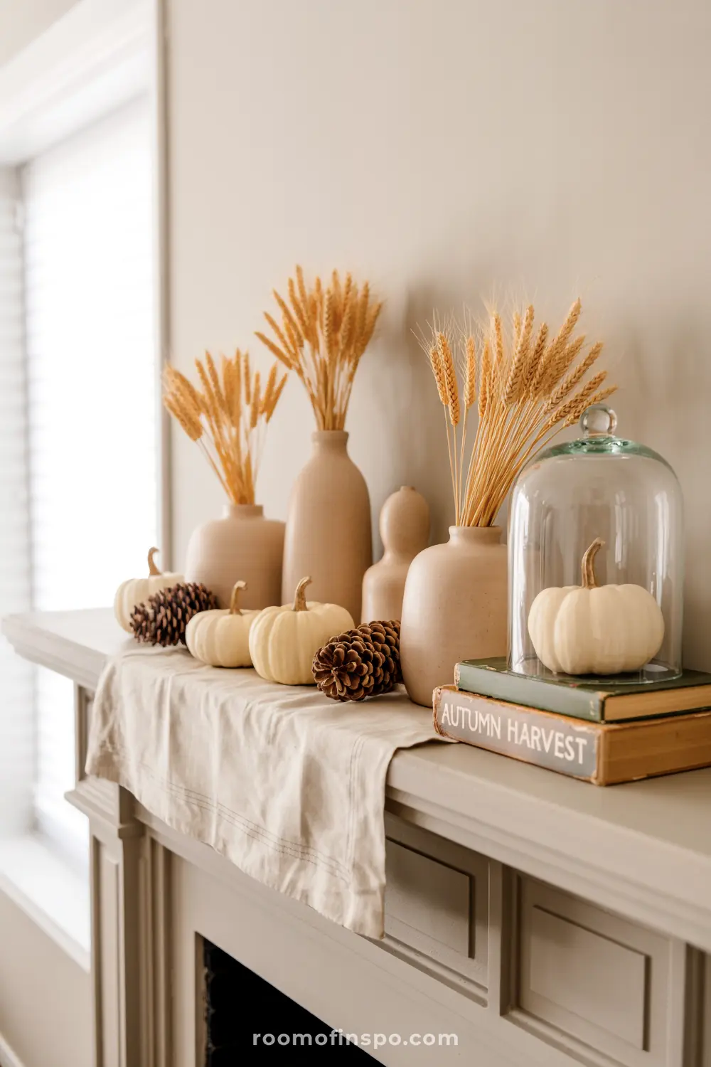 A tasteful fall mantel features white pumpkins, pinecones, and wheat stalks in vases, with a cloche over a pumpkin and "Autumn Harvest" books.