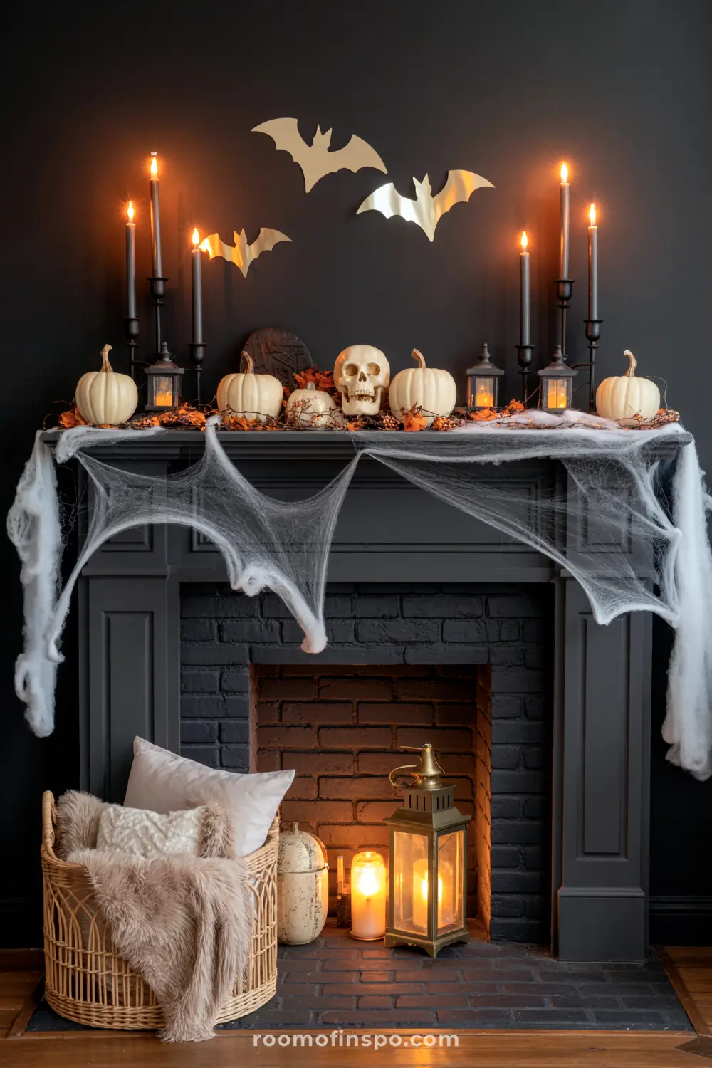 A striking Halloween mantel on a dark fireplace features white pumpkins, a skull, candles, lanterns, and eerie cobwebs, with bats on the wall.