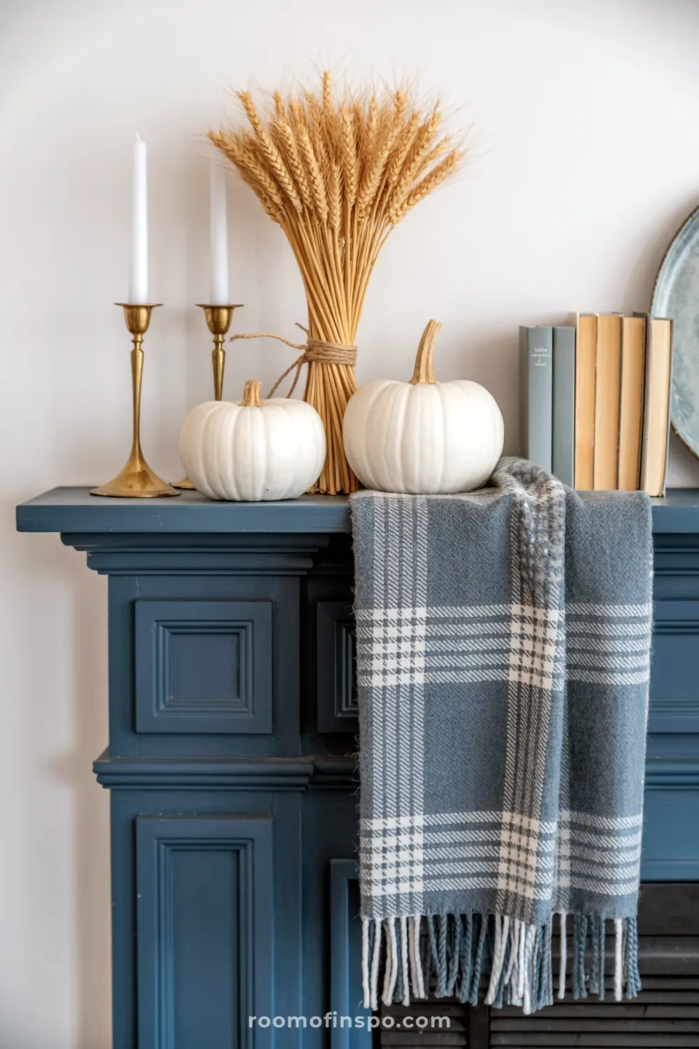 A stylish blue mantel is decorated for fall with white pumpkins, a bundle of wheat, brass candlesticks, and a cozy plaid throw.