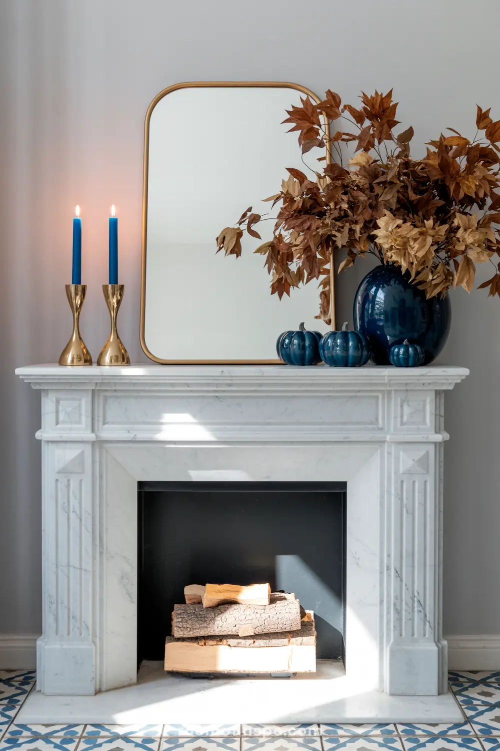 A elegant fall mantel features a gold-framed mirror, blue pumpkins, and a vase with dried autumn leaves, alongside blue candles in brass holders.