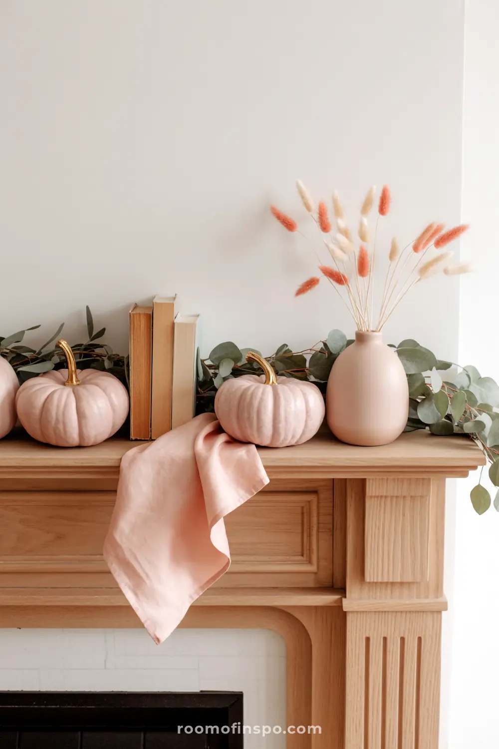 A soft blush-toned fall mantel features decorative pumpkins, dried grasses in a vase, and a eucalyptus garland, creating a serene autumn display.