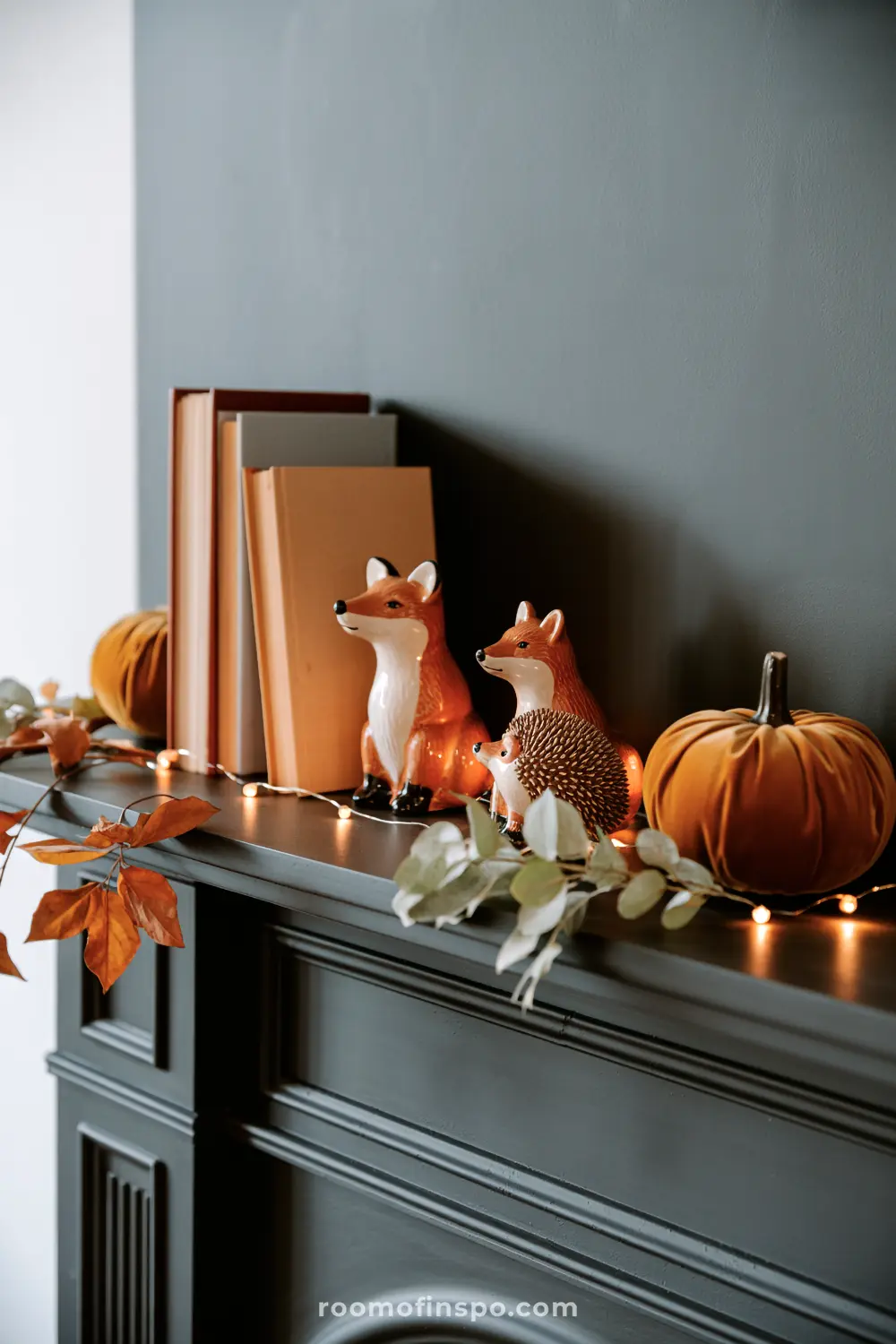 A fireplace mantel is adorned with charming fall home decor, including fox figurines, velvet pumpkins, books, and delicate autumn fairy lights.