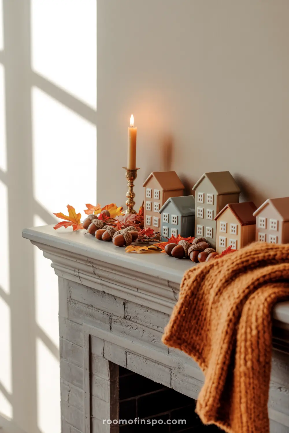 A quaint fall mantel features miniature houses, golden acorns, autumn leaves, and a lit candle, alongside a cozy knitted throw.