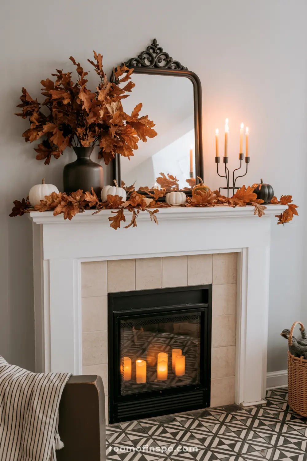 A cozy fireplace mantel features elegant fall home decor with a vase of brown leaves, white pumpkins, candles, and an autumn leaf garland.