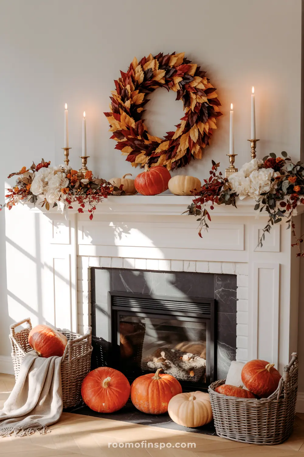 A stunning autumn mantel features a vibrant leaf wreath, white and orange pumpkins, floral garlands, and tall candles, with more pumpkins in baskets below.