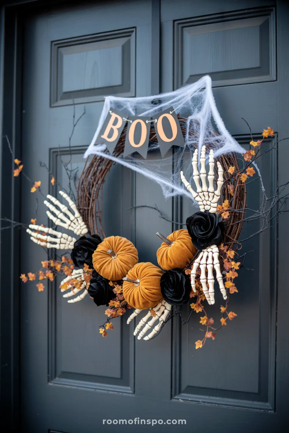 A spooky fall wreath with velvet pumpkins, skeleton hands, and a "BOO" banner hangs on a dark gray front door for Halloween.