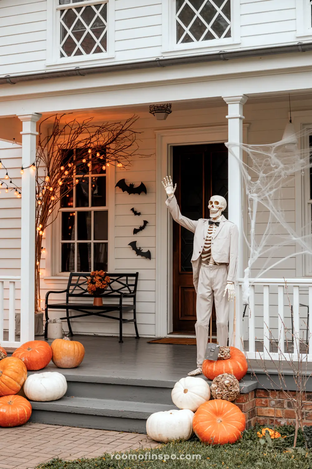 A Halloween front porch is decorated with a dapper skeleton, bats, spiderwebs, bare branches with lights, and various pumpkins on the steps.