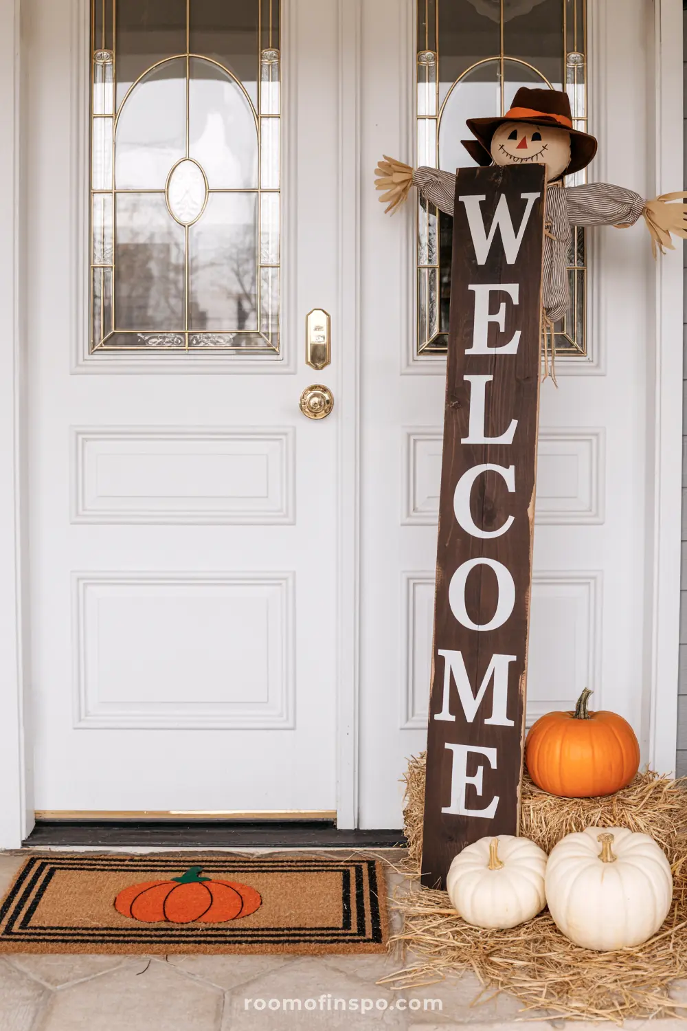 A friendly scarecrow and "WELCOME" sign adorn a fall front porch, with pumpkins and a pumpkin mat for autumn.