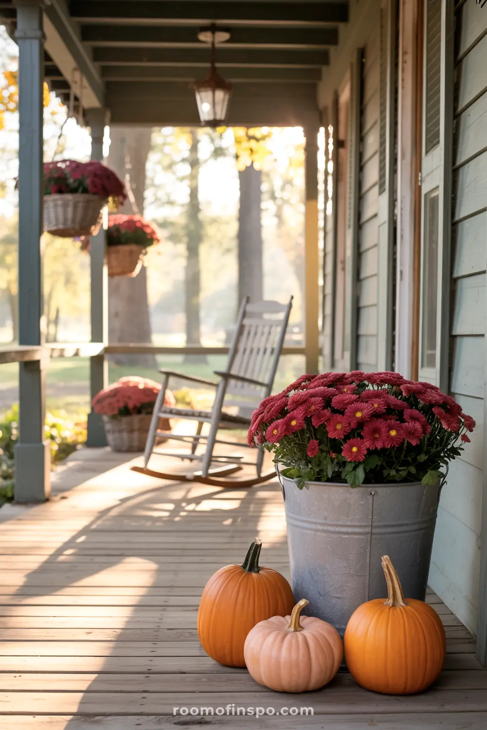 Potted red mums and pumpkins adorn a rustic fall front porch, inviting relaxation on a bright autumn day.