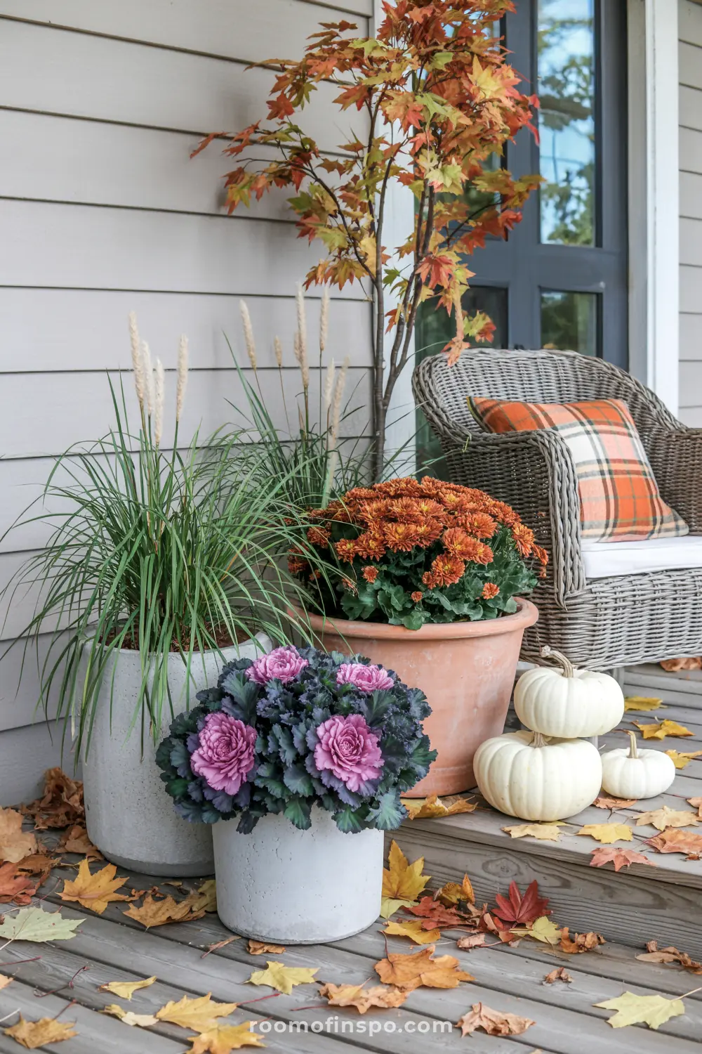 Potted mums, ornamental kale, and pumpkins create a vibrant fall front porch display with autumn leaves scattered.