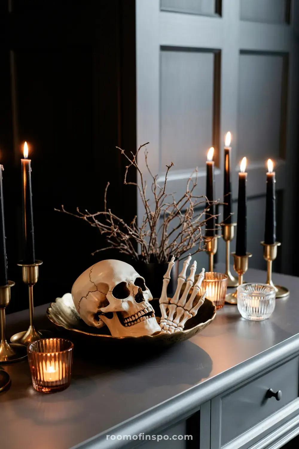 A macabre Halloween centerpiece showcasing a skull and skeletal hand in a golden bowl, flanked by lit black candles and dry branches.