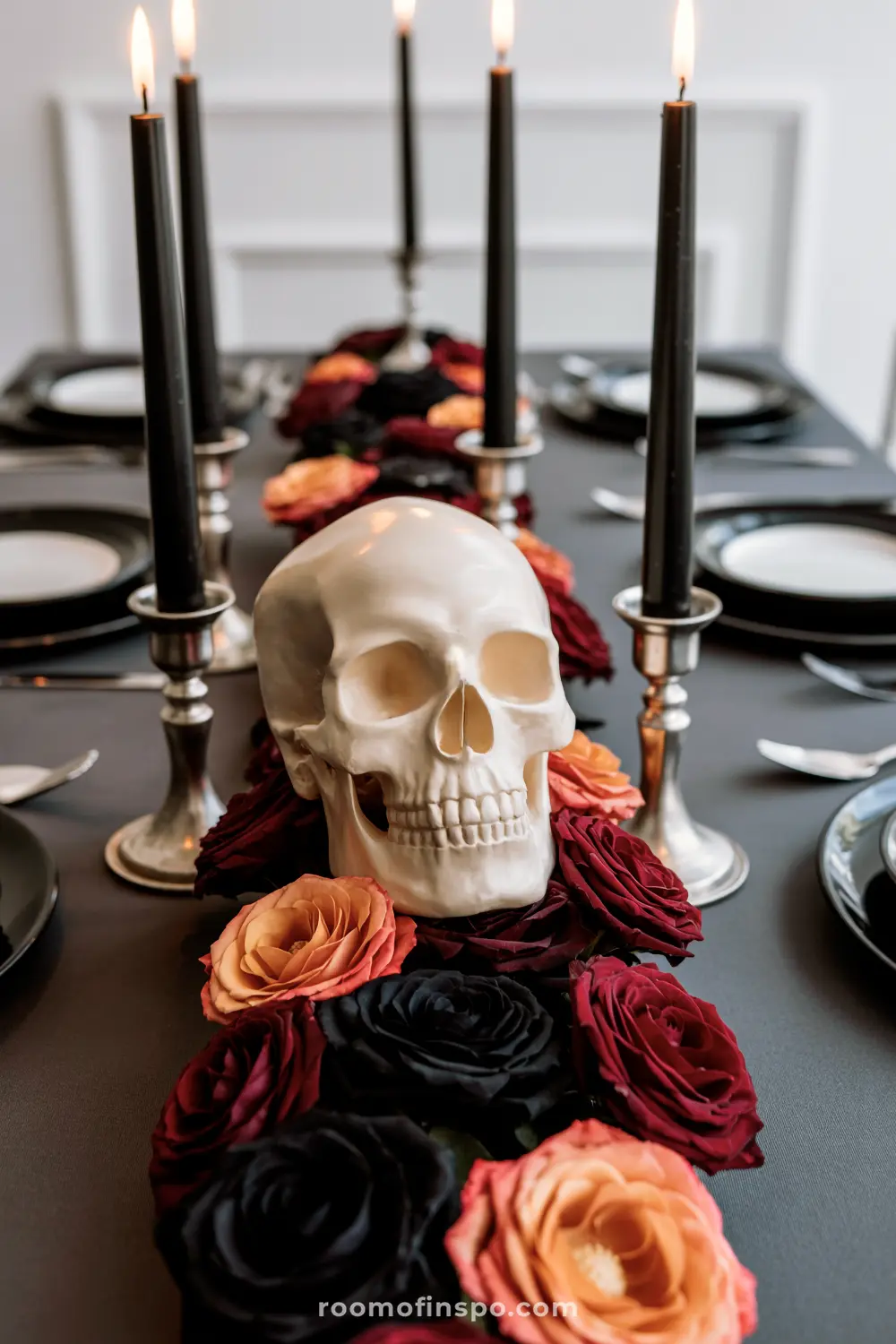 A sophisticated Halloween centerpiece features a skull surrounded by a lush runner of black, red, and orange roses, with tall black candles.