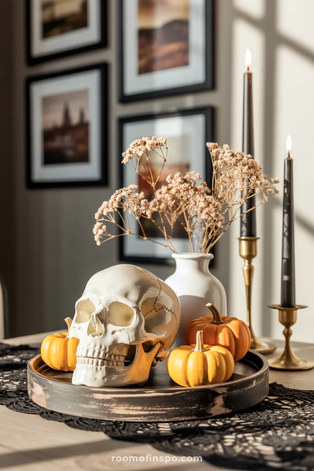 A sunlit Halloween centerpiece featuring a skull, mini pumpkins, and dried flowers in a white vase on a rustic tray.